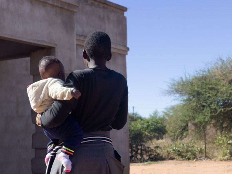 A young woman holding a baby outside a house, photographed from behind to protect their identity